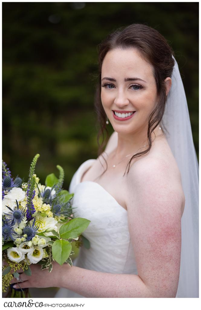 bridal portrait of a bride wearing a white sleeveless dress, and a white veil, her bouquet is white, green, and purple, she's smiling and standing with her side to the camera