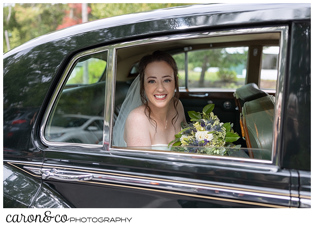 a bride wearing a white dress and veil, with a yellow, white, green, and purple bouquet, smiles from the back of a vintage Bentley