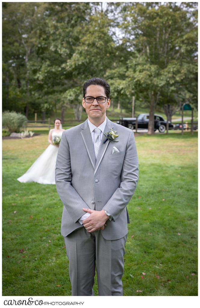 a groom wearing a gray suit, stands with his back to his bride, during their wedding day first look