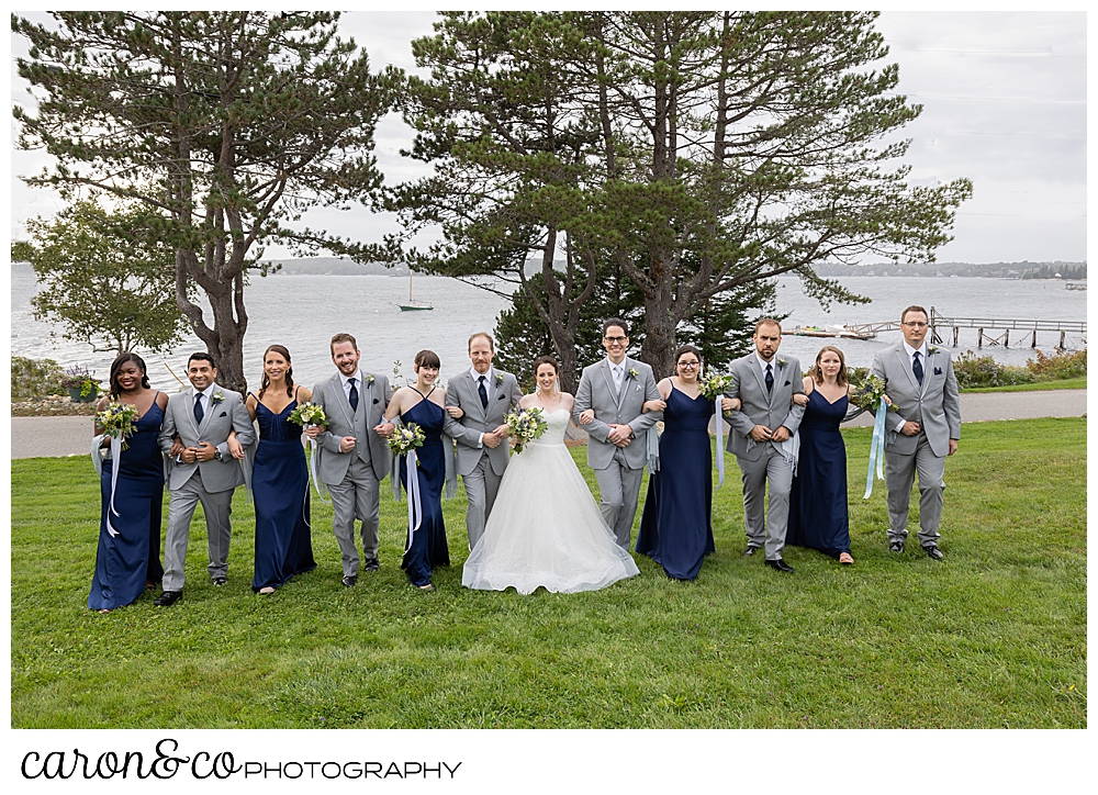 a bridal party of groom and groomsmen in gray, bridesmaids in navy, and bride in white, link arms and walk across the front lawn at th Spruce Point Inn, Boothbay Harbor, Maine