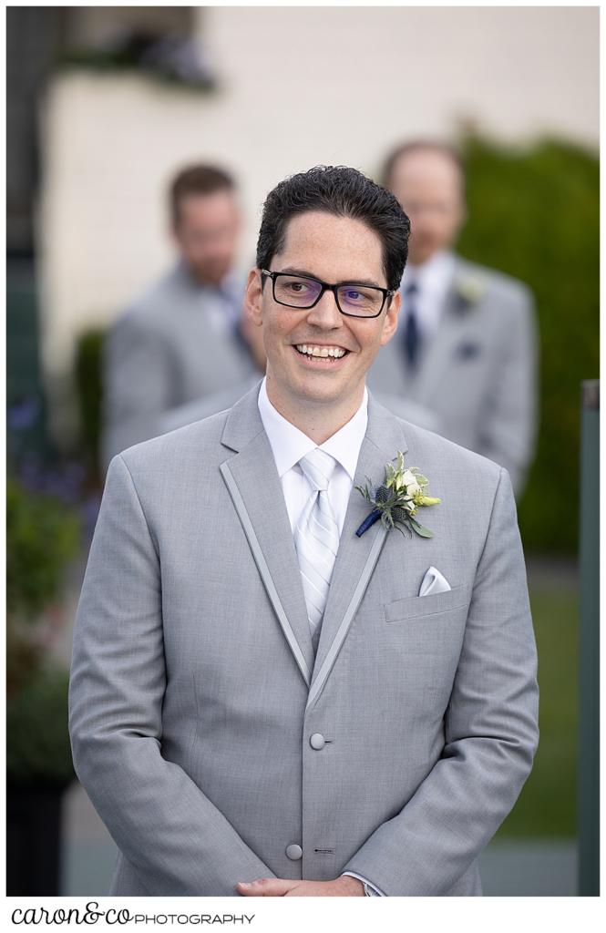 a smiling groom in a gray suit, walks towards the ceremony on the deck at the Spruce Point Inn, Boothbay Harbor, Maine