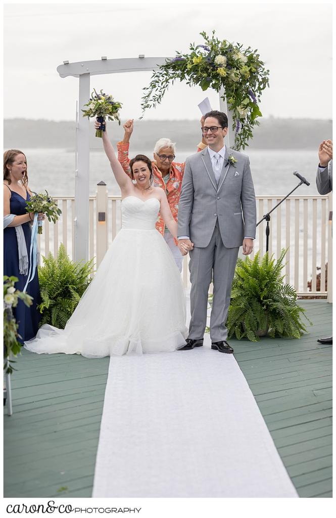 a bride, holding her bouquet aloft, and groom smile as they process down the aisle after their Spruce Point Inn Maine wedding ceremony