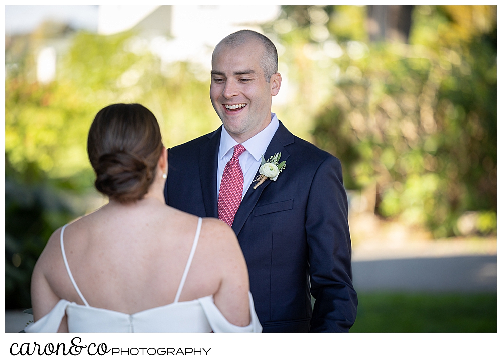 a groom wearing a blue suit with a red tie, smiles as he walks towards his bride during a wedding day first look