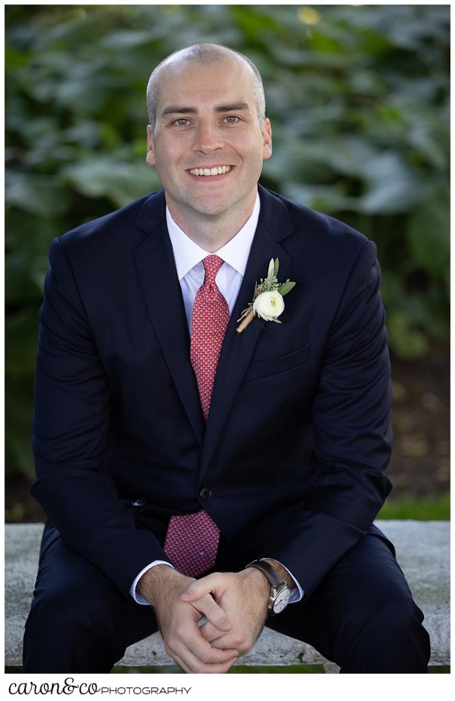 portrait of a groom wearing a navy suit, sitting on a bench, he is leaning forward with his hands clasped