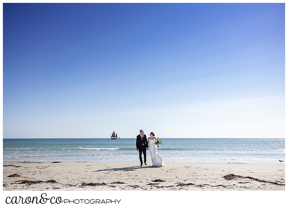 a bride and groom walk on Gooch's Beach in Kennebunk, Maine, a sailboat sails in the distance
