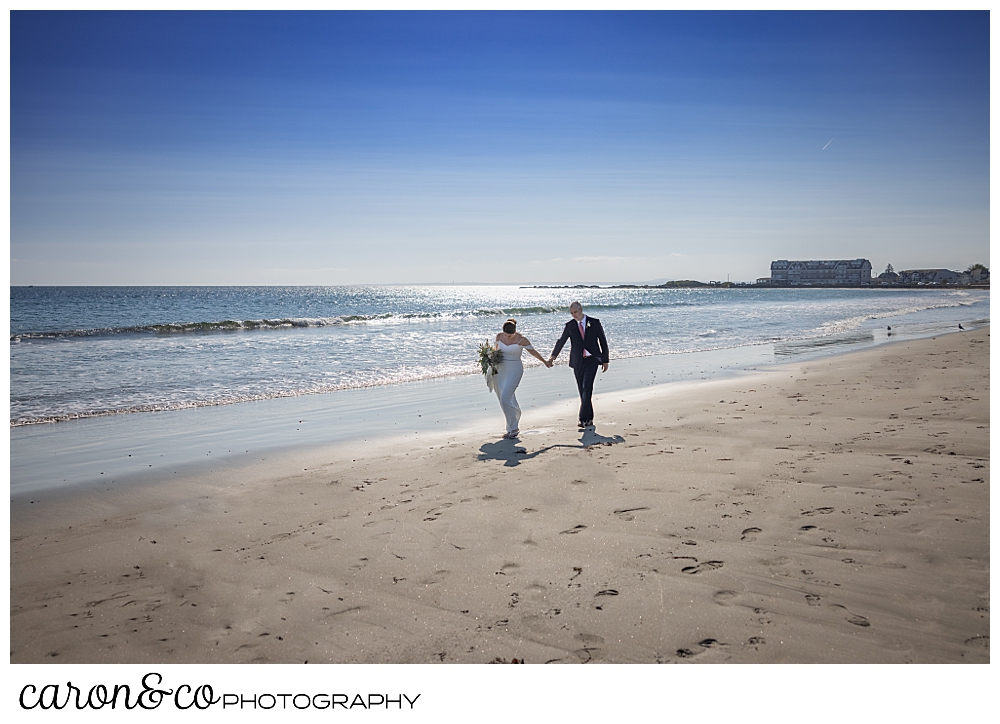 a bride and groom hold hands and walk along the water on Gooch's Beach, Kennebunk, Maine