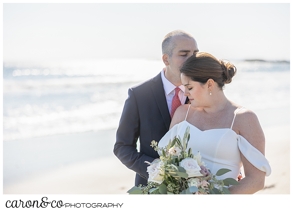 a groom stands behind his bride, kissing her temple, the bride has her eyes closed, and holds a bouquet of pink and green