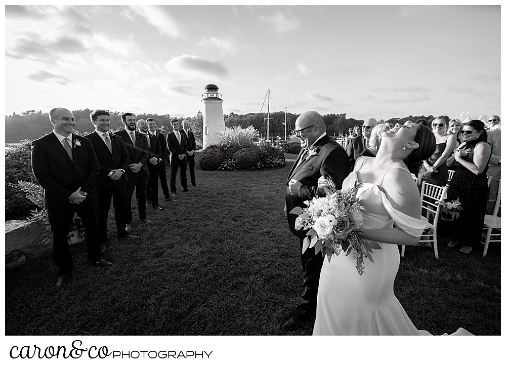 a black and white photo of a bride and her dad walking towards the groom at the outdoor ceremony site at a Nonantum Resort Kennebunkport Maine wedding. The bride has thrown her head back as her dad has begun to cry