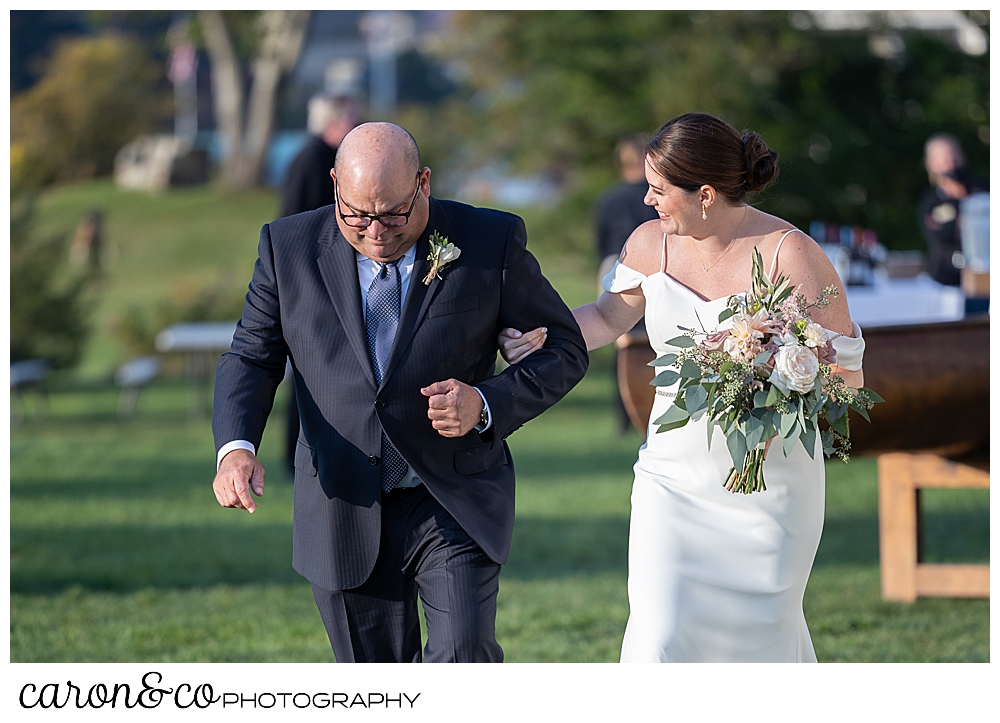 a bride and her father begin their walk down the aisle at the outdoor ceremony location at The Nonantum Resort, Kennebunkport, Maine, the bride smiles as she looks at her dad, as he looks down at the ground