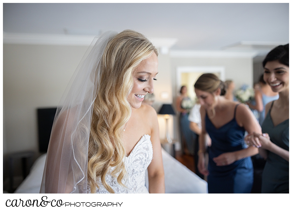 a bride wearing a white dress and veil, smiles as her bridesmaids in the background help with her dress