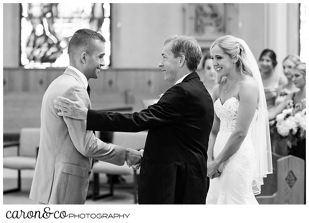 a black and white photo of a father of the bride shaking hands with the groom as the bride looks on at St. Joseph's Church Biddeford Maine