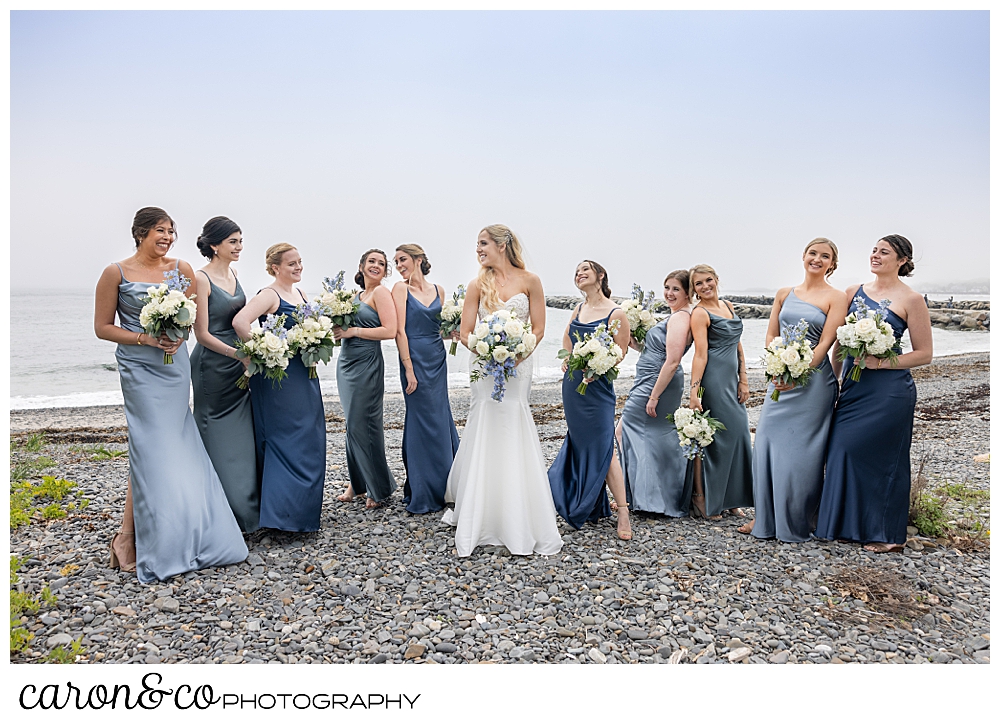 a bride stands in the middle of her bridesmaids who are dressed in different tonal blue dresses, on Colony Beach Kennebunkport Maine
