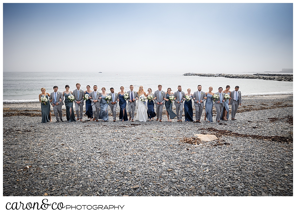 a large bridal party, the women in shades of blue, the men in gray, walk arm in arm on Colony Beach, Kennebunkport, Maine
