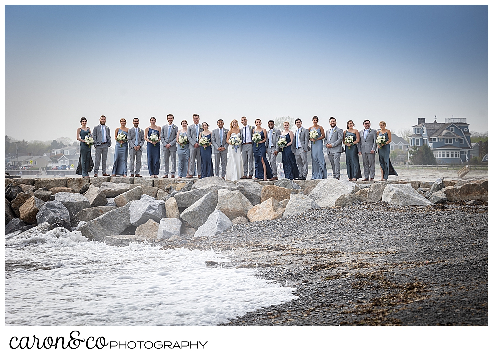 a large wedding party wearing blue and gray, stand together on the Kennebunkport Breakwater, Kennebunkport Maine