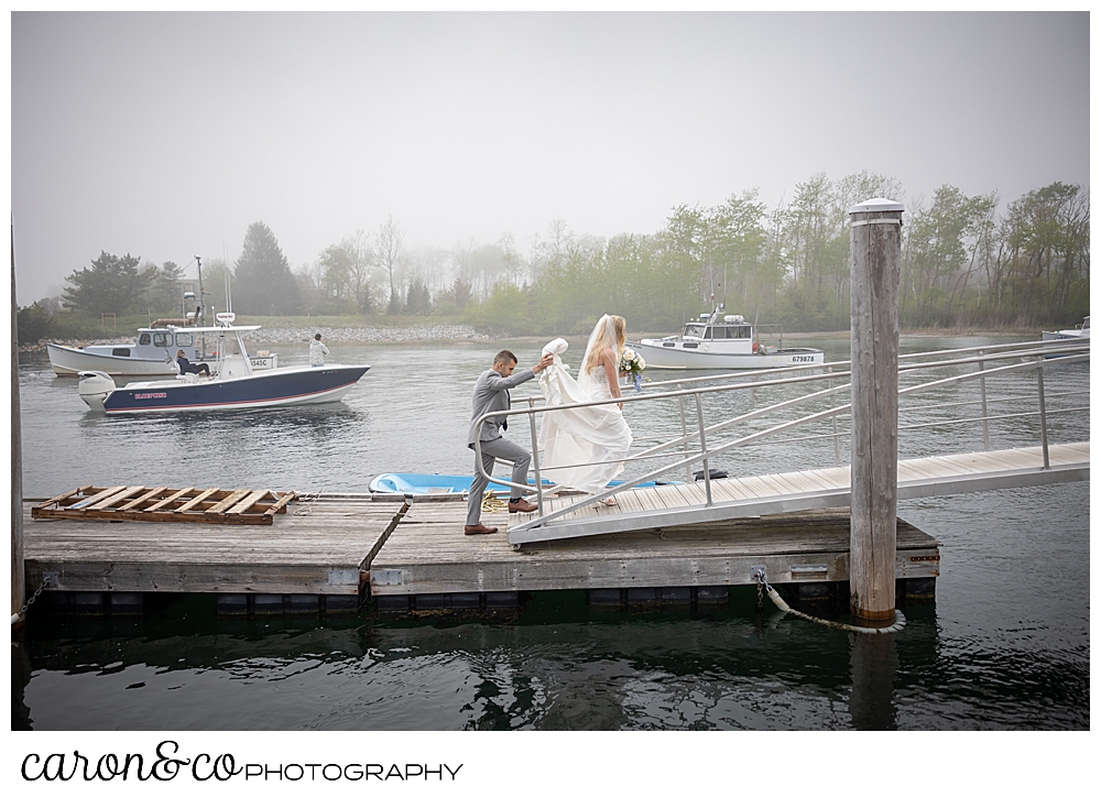 a bride is walking on a dock, her groom is helping with her train during a breakwater inn Kennebunkport wedding, Kennebunkport, Maine