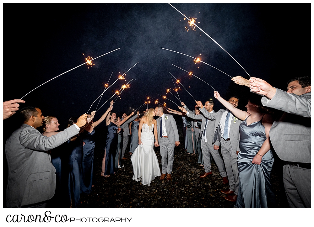 a bride and groom are kissing as their wedding guests hold sparklers in an arch over them, on a beach in Kennebunkport, Maine, during a breakwater in Kennebunkport wedding reception