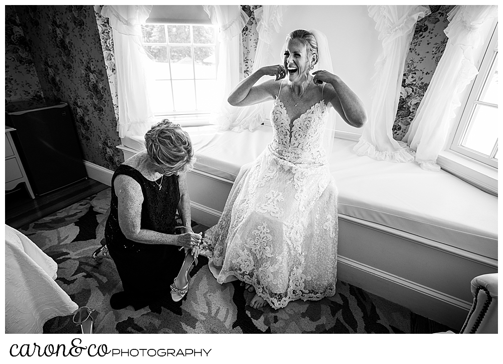black and white photo of a bride laughing as she adjusts her dress, her mother is helping her put her shoes on