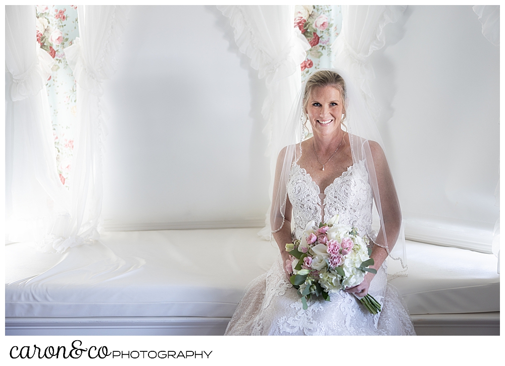 a beautiful bride sits on a window seat, holding her bridal bouquet, and smiling