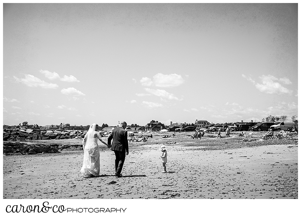 black and white photo of a bride and groom walking on colony beach, there's a little girl standing near them, Kennebunkport Maine