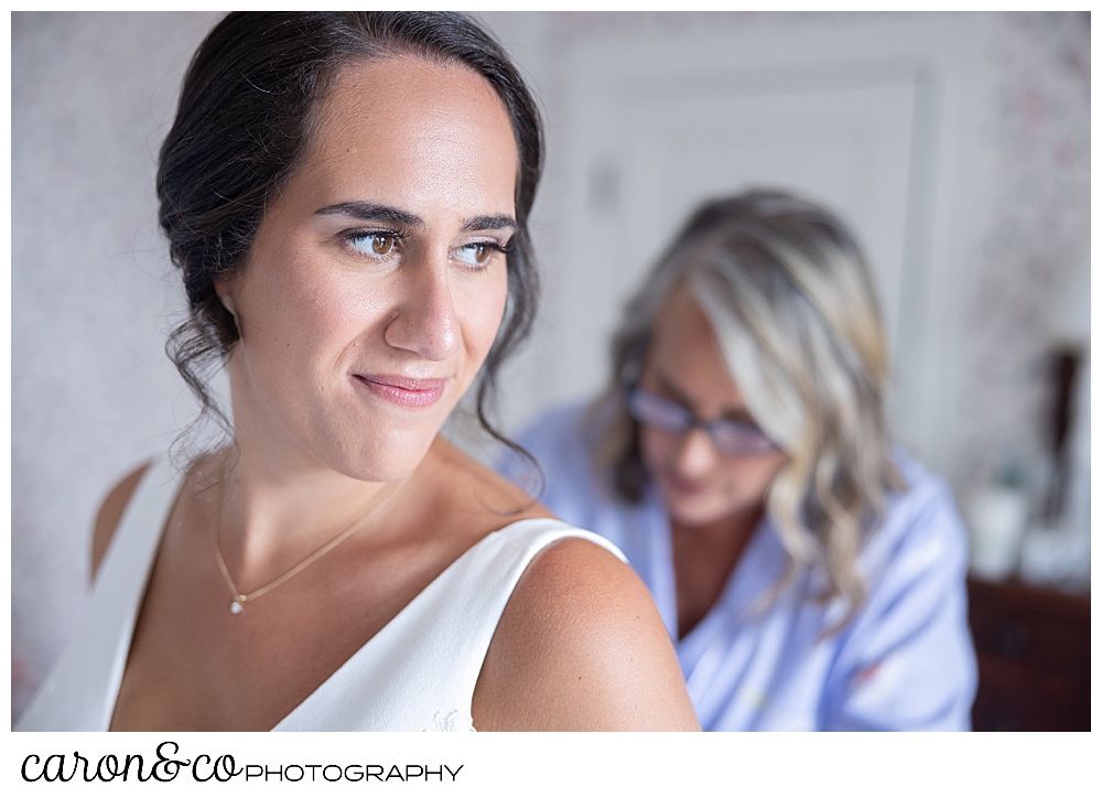 a bride in the foreground smiles, as her mom in the background, helps her get into her wedding dress