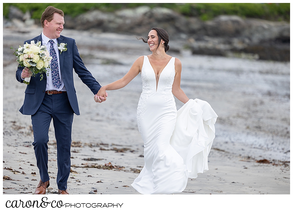 a bride and groom walk on the beach, holding hands, looking at each other, the bride holds her dress, and the groom holds the bridal bouquet