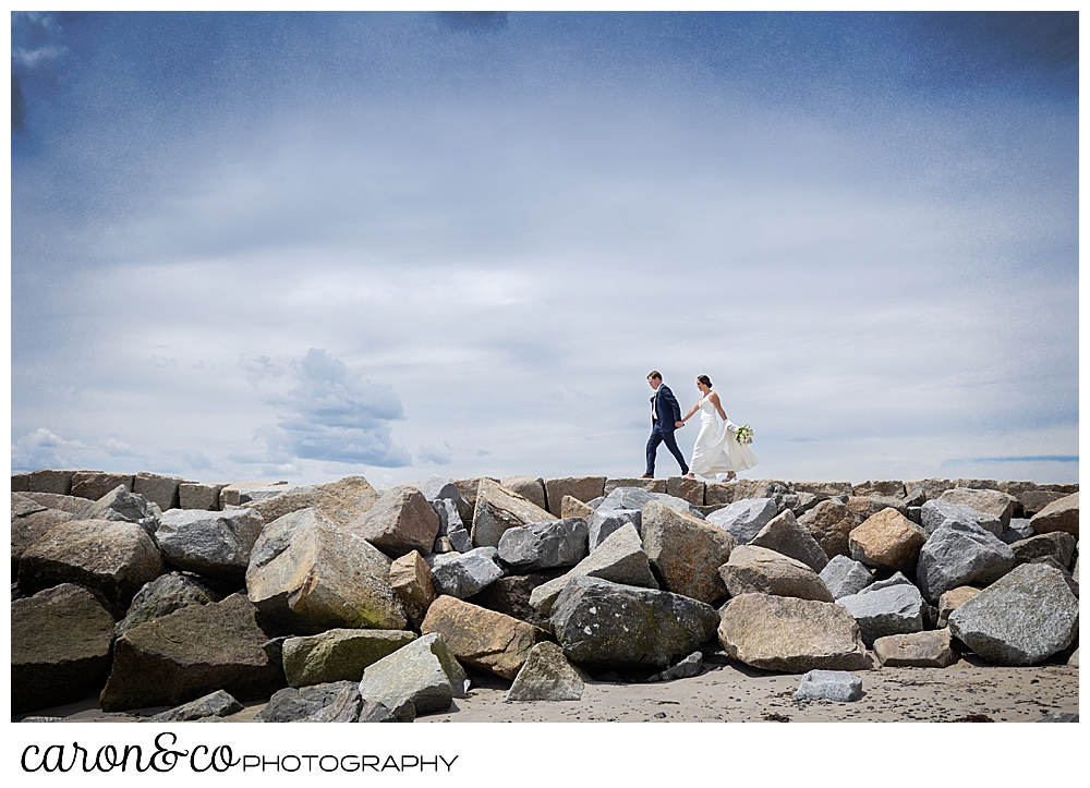 a bride and groom walk on the Kennebunkport Maine breakwater, the groom walks in front, holding the brides hand as she walks behind