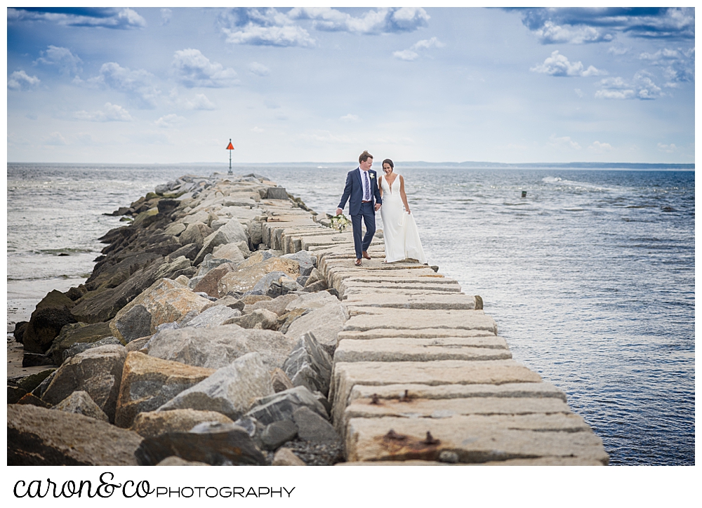 a bride and groom walk side by side, holding hands, on the Kennebunkport Maine breakwater at Colony Beach