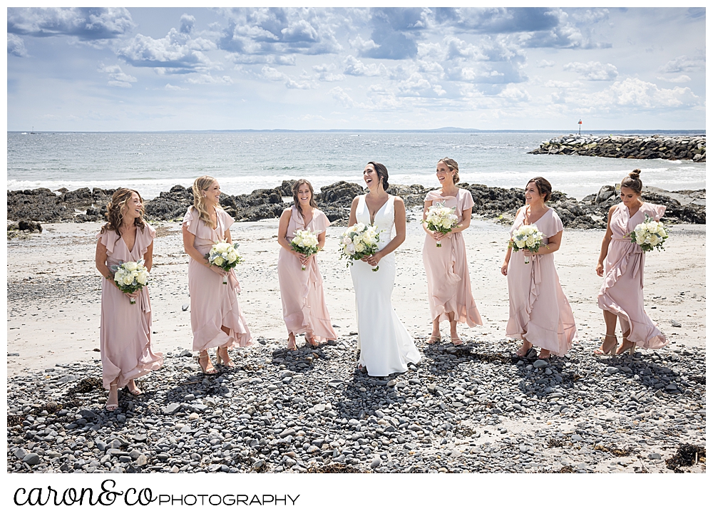 a bride is standing on Colony Beach in Kennebunkport, Maine, with her bridesmaids who are wearing pastel pink dresses, carrying white and green bouquets