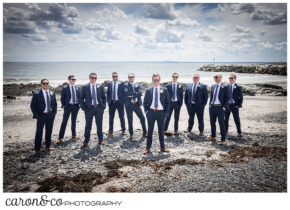 a groom and his groomsmen, wearing blue suits and sunglasses, stand on colony beach in Kennebunkport Maine