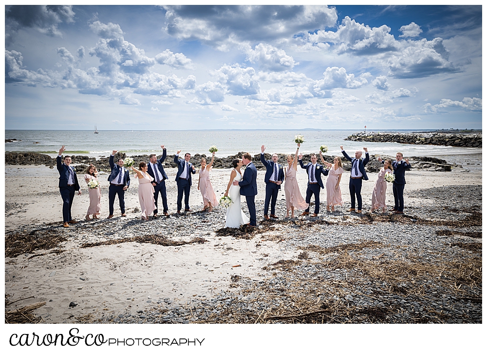a bridal party, with the bridesmaids wearing pink, the bride wearing white, and the groom and groomsmen wearing blue, cheer while the bride and groom kiss, on Colony Beach in Kennebunkport Maine