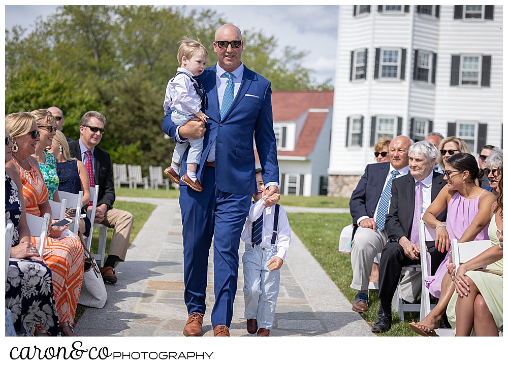 a groomsman in a blue suit, walks down the outdoor aisle holding a young boy, and holding the hand of an older boy, during a Kennebunkport Maine wedding at the colony hotel