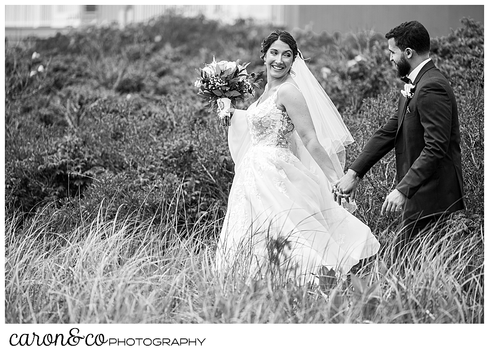 a black and white photo of a bride and grooming, holding hands and walking, the bride is slightly in front of the groom, and looking back at him