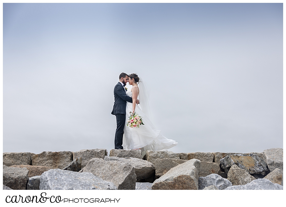 a bride and groom stand face to face, their foreheads touching, on the Kennebunkport Maine breakwater