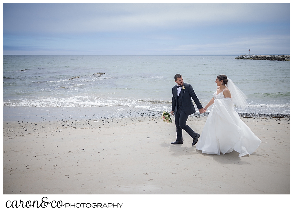 a bride and groom are walking on Colony Beach, Kennebunkport, Maine, the groom is ahead of the bride, and looking back at her