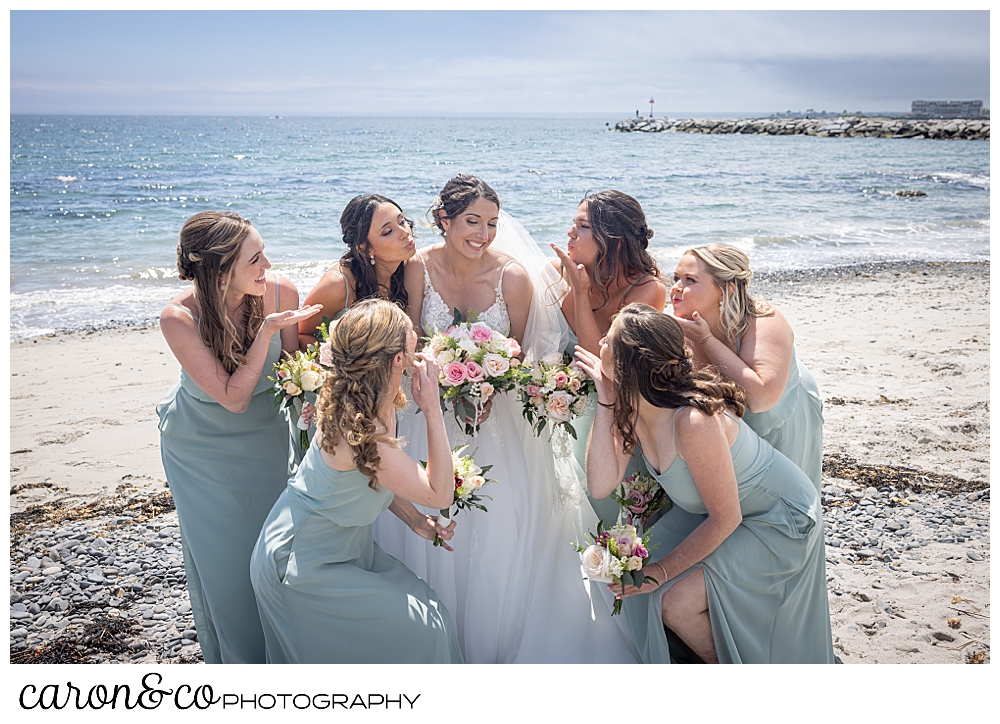 a bride is surrounded by her bridesmaids, who are blowing kisses, on Colony Beach, Kennebunkport, Maine