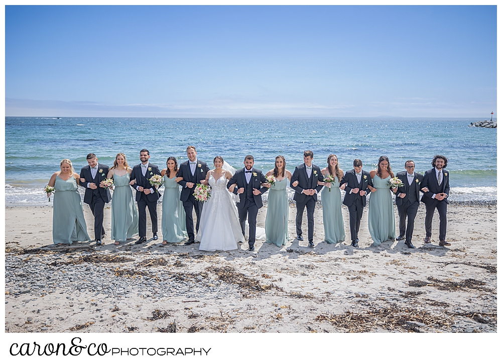 a bride and groom with their bridal party, walk arm in arm on Colony Beach, Kennebunkport, Maine