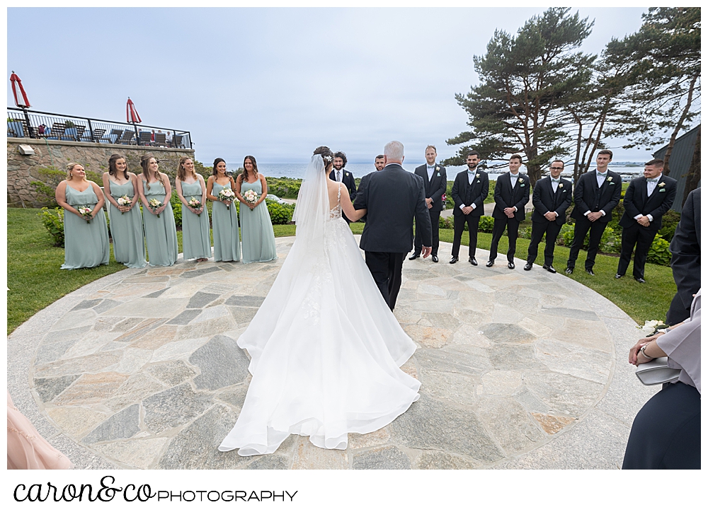 A bride and her father, from the back, approach the groom, the bridal party are on either side, at a spring wedding at the colony hotel Kennebunkport Maine