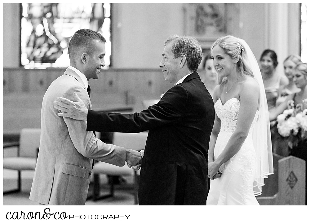 a black and white photo of a bride smiling as her dad and groom shake hands at a St. Joseph's Biddeford Maine wedding ceremony