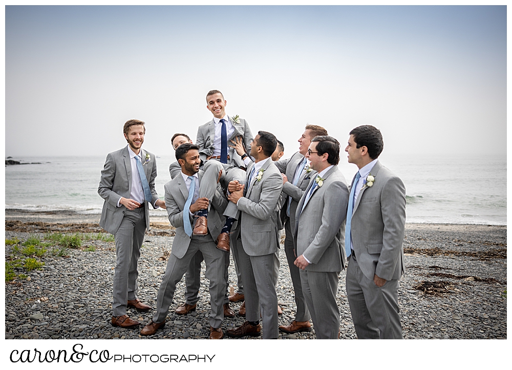 a groom is held aloft by his groomsmen, all wearing gray suits, at Colony Beach, Kennebunkport, Maine